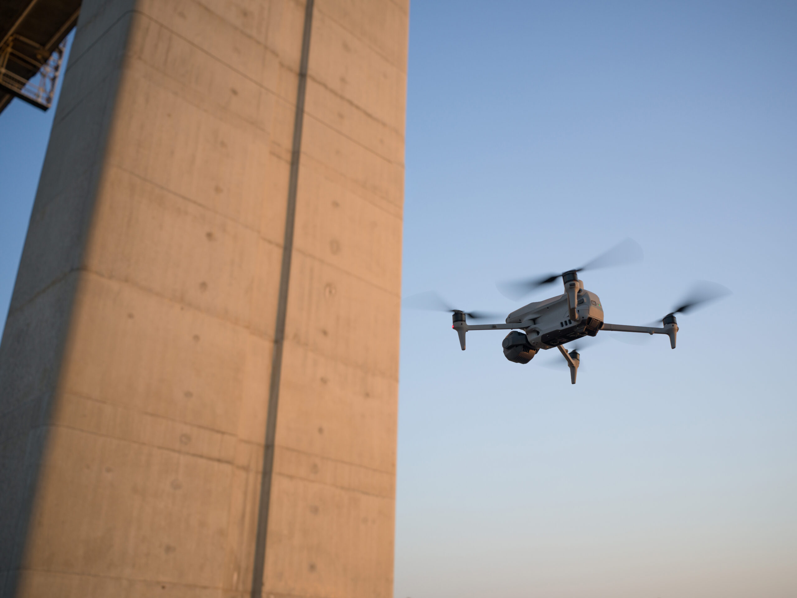 Inspektionsdrohne fliegt im Abendlicht dicht an einem Brückenpfeiler aus Beton entlang