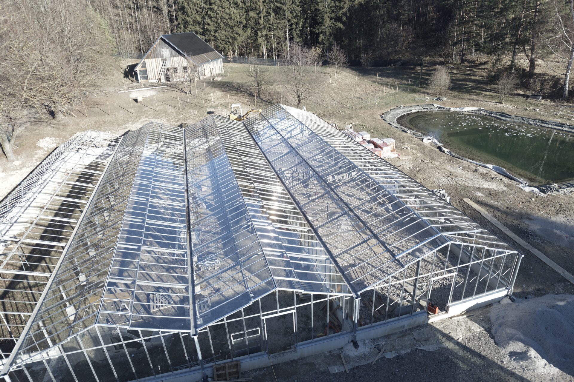 Luftaufnahme eines im Bau befindlichen Glashauses mit Baumaterial und Teich auf einer Baustelle
